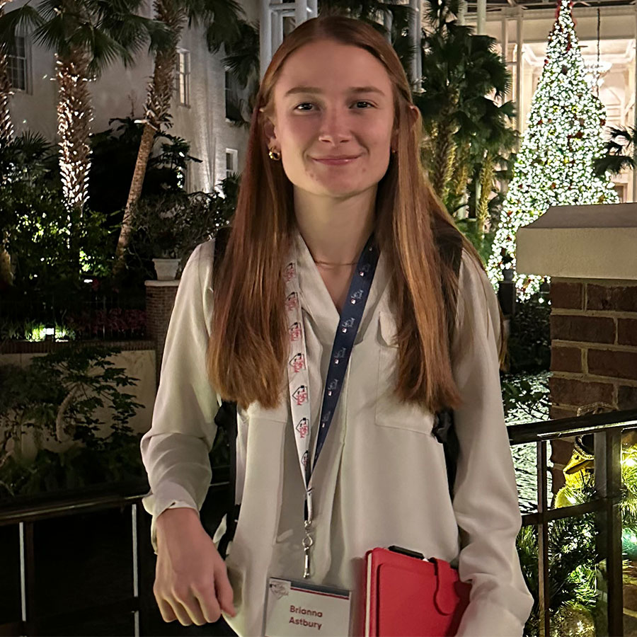 A student with long brown hair, wears a white blouse with a lanyard around her neck, smiling at the camera in a tropical location decorated for the winter holidays.