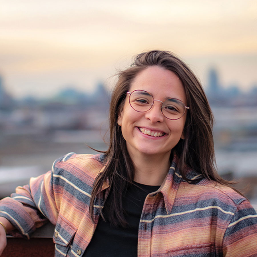 A woman with shoulder-length brown hair and glasses smiles at the camera with a city skyline in the distance.