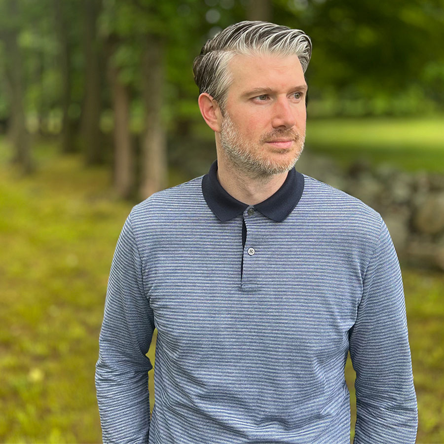 A man in a striped, collared grey shirt looks into the distance while outdoors.