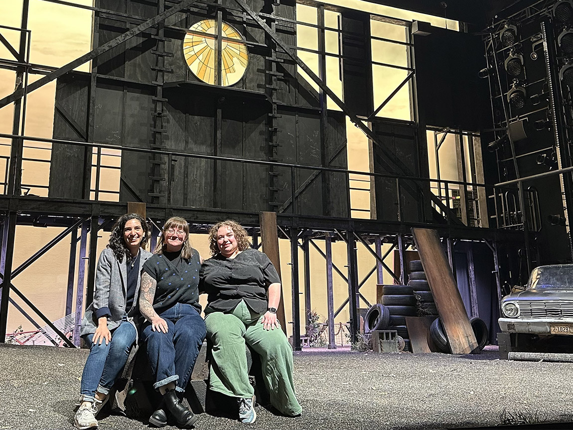 Three alumni sit on the set of The Outsiders — a large theatrical flat, viewed from the back. It looks like an abandoned barn, with light shining through several windows. A dilapidated 1960s car sits all the way to the right.