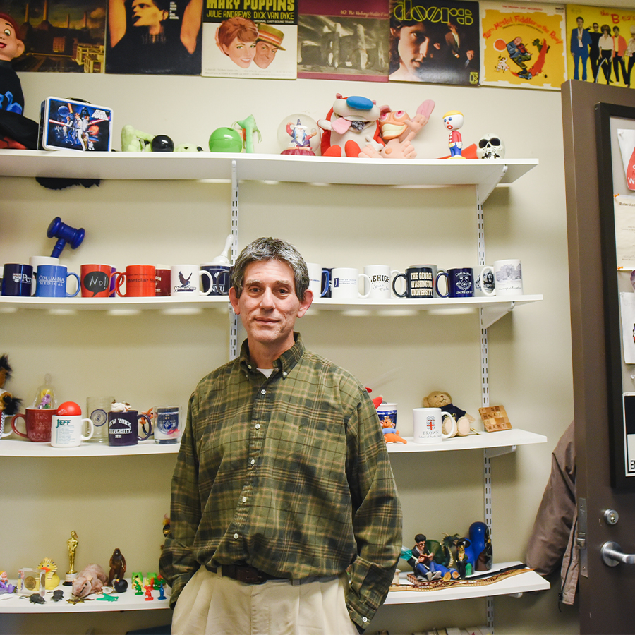 A man in a green plaid shirt stands, arms in pocket, in front of a shelf of colorful knick knacks and art.