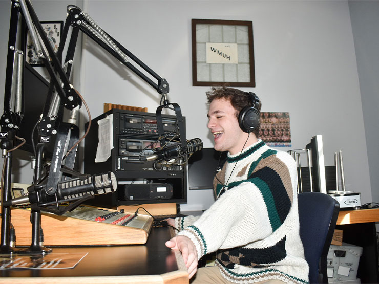A male student in a sweater sits speaks into a microphone in a radio studio.