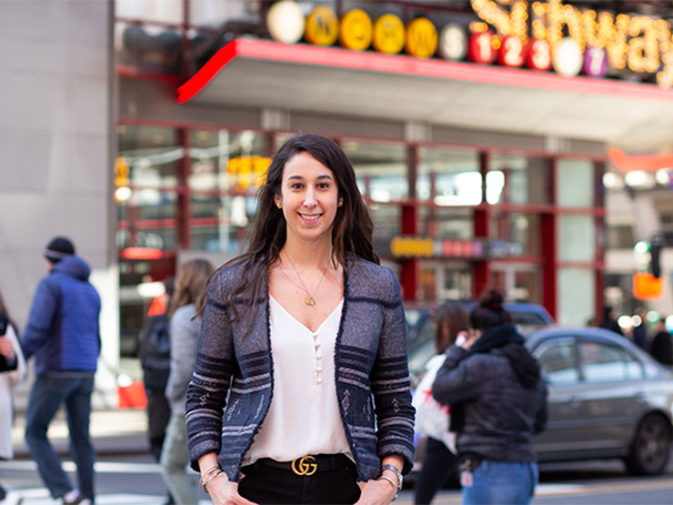 An adult with long brown hair, a white blouse and grey and black blazer smiles at the camera while standing at a busy city intersection.