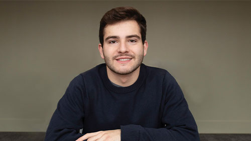 A male student in a dark blue crew neck sweatshirt smiles at the camera.
