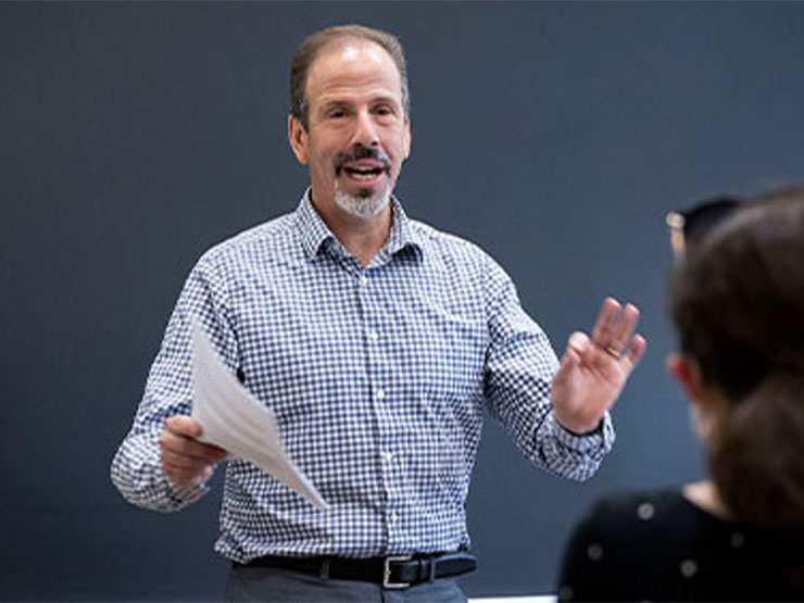 A college professor speaks to his students at the front of a classroom in front of a chalkboard.