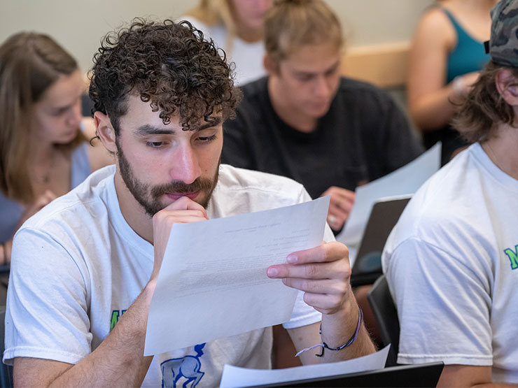 A student studies a piece of paper, surrounded by classmates at desks.