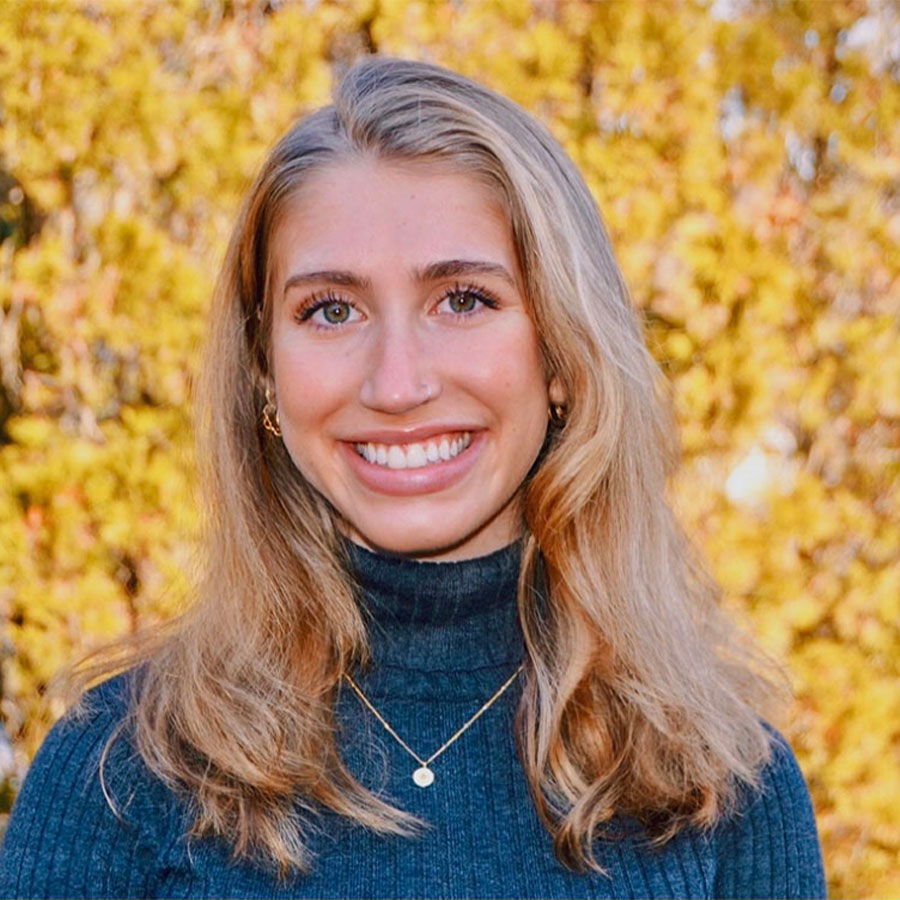 An adult with long blonde hair wears a dark blue turtleneck sweater while smiling at the camera outdoors on an autumn day.