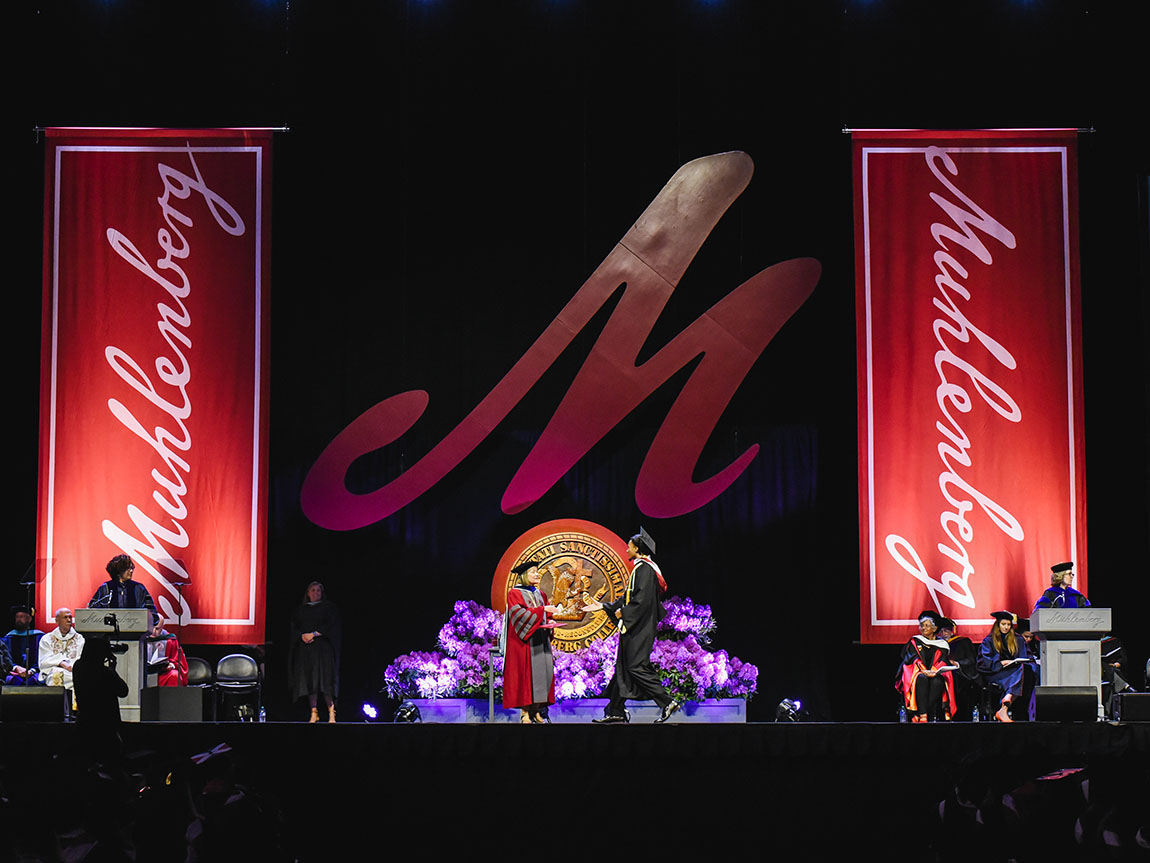 A student receives their college diploma on a stage that has a big red M hanging behind it