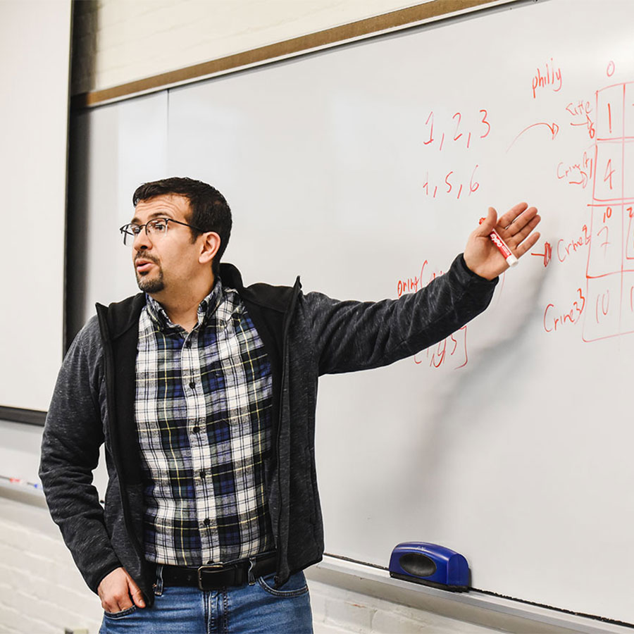 An instructor in a plaid shirt and grey jacket speaks to a class while pointing at formulas scrawled on a whiteboard.