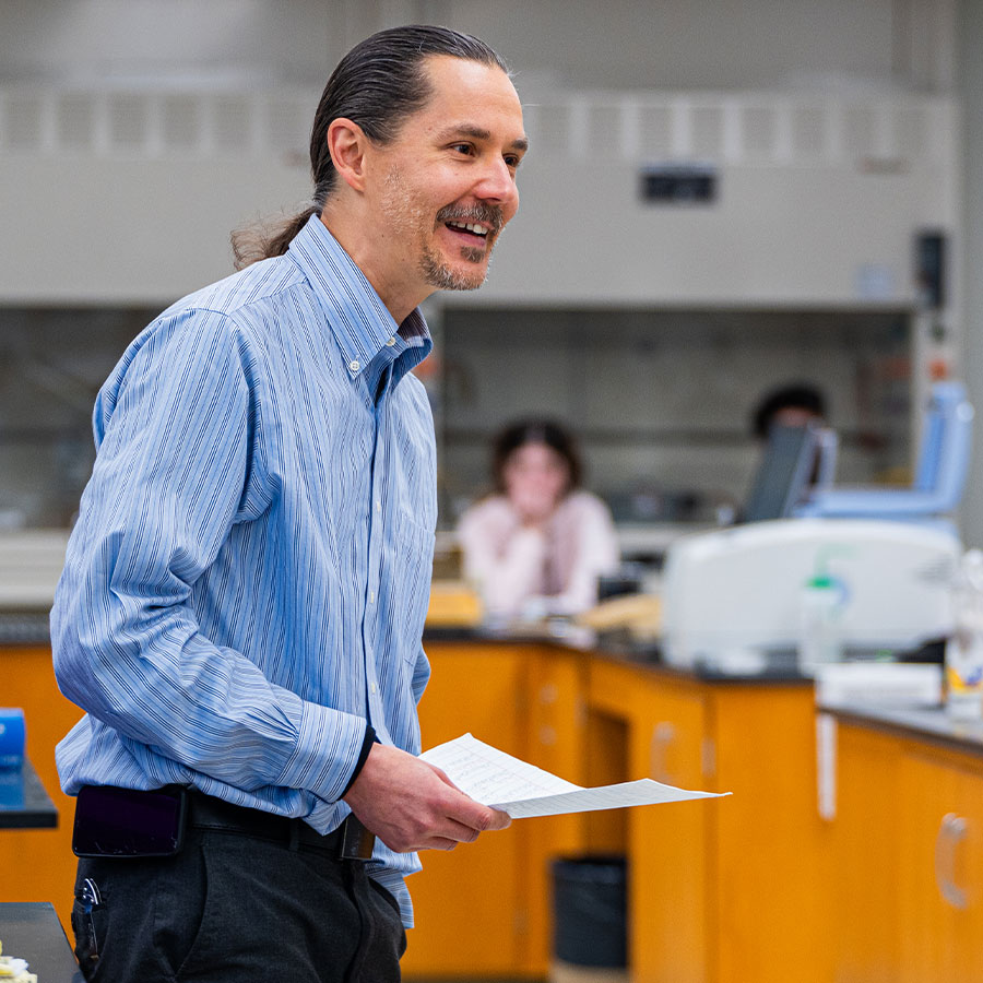 An adult instructor in a blue dress shirt and long ponytail smiles while speaking to students in a lab.