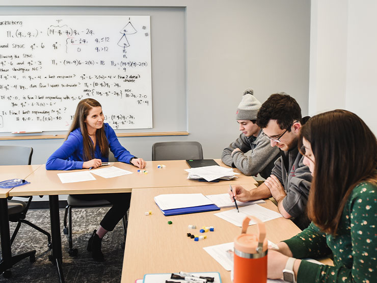 An instructor in a bright blue blouse and long brown hair addresses a table full of students in a classroom with complicated formulas written across a whiteboard.