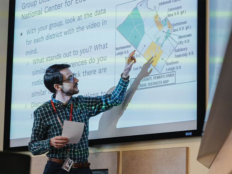 An adult instructor in a plaid shirt stands in front of a projector screen and points to a map while speaking in front of a classroom.