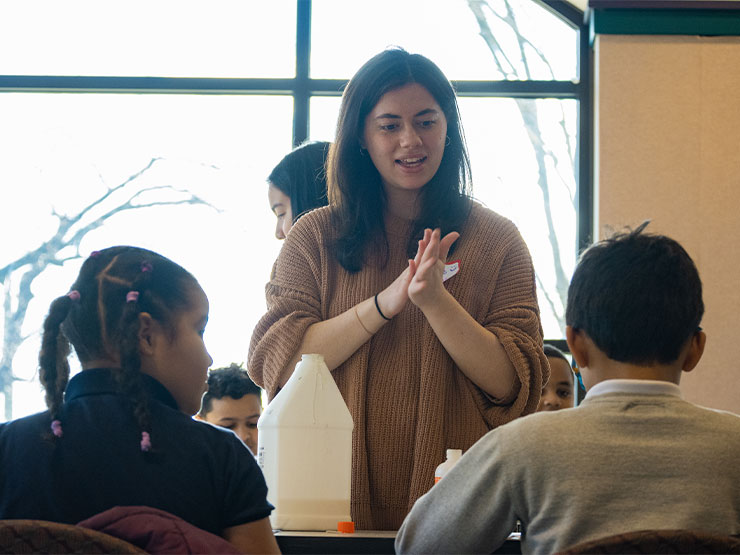 A young adult in a sweater speaks to elementary school children in a room with tables stocked with crafting materials.