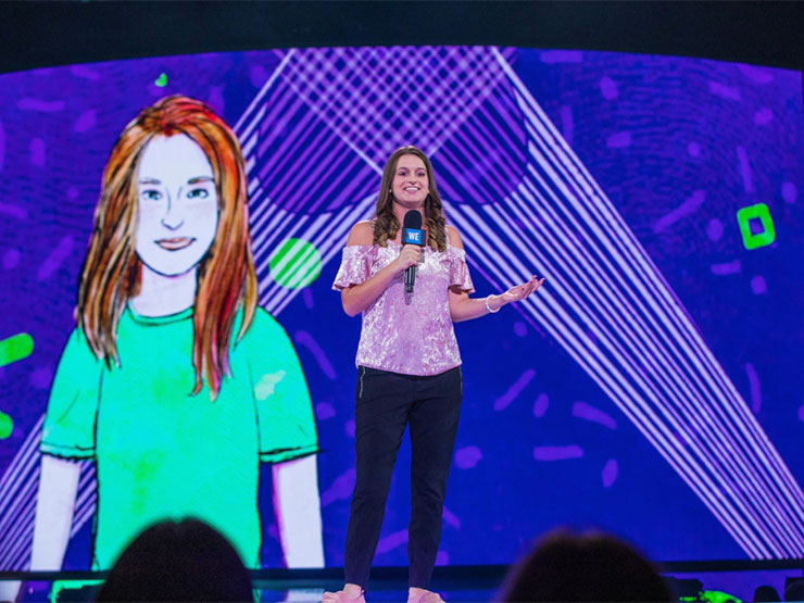 A young adult speaks on a stage, holding a microphone, with an illustration pictured on screen behind them.