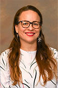 Assistant professor Lilianne Herrera wearing white shirt and glasses posed for portrait in front of beige abstract background.