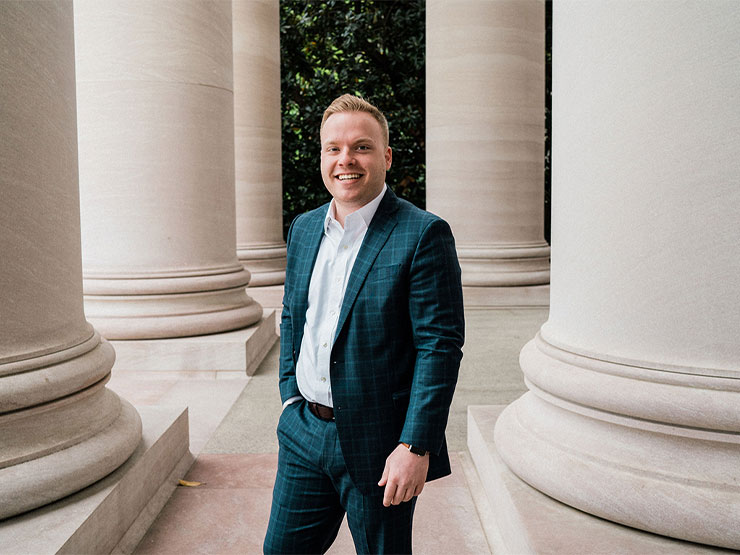 A man in a dark blue suit and smiles, one hand in pocket, outdoors near large marble columns.