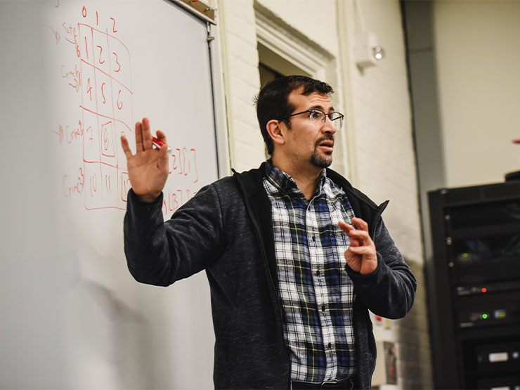 An instructor in a flannel shirt and black pullover speaks to a class and points to figures written on a white board.