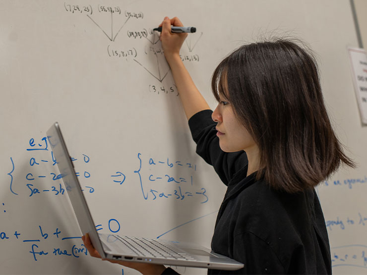 A student holds an open laptop with one hand and writes formulas on a whiteboard with the other.