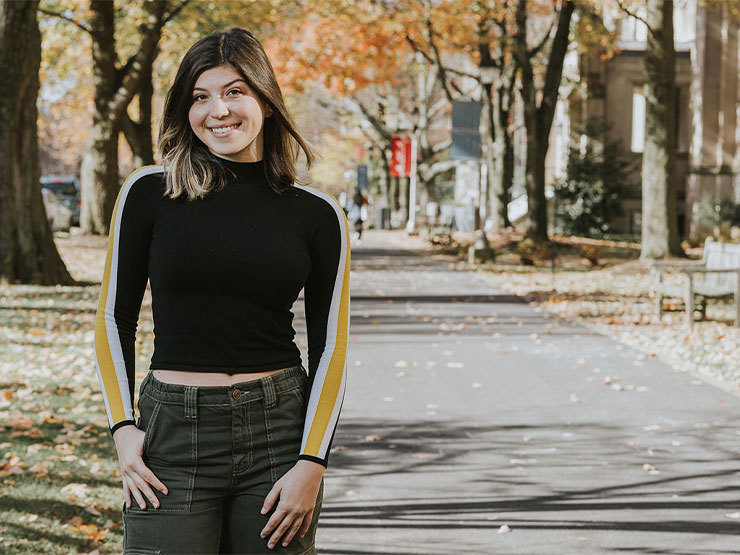 A young adult woman smiles at the camera while standing on a college campus during autumn.