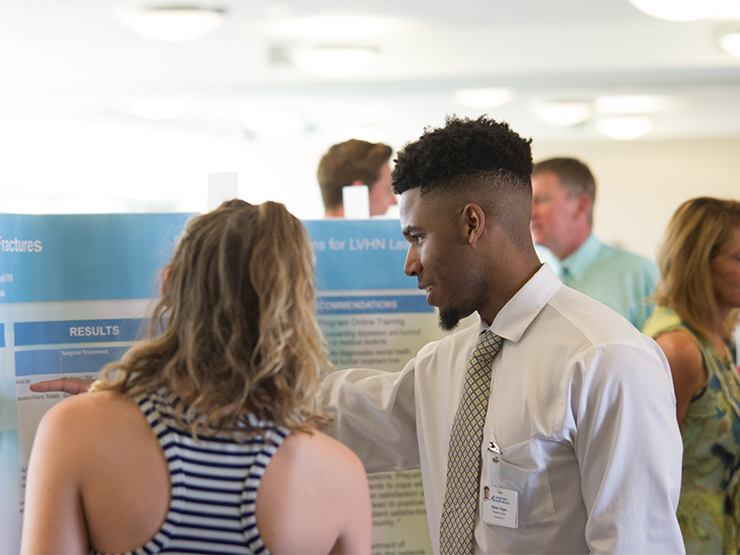 A student in a dress shirt and tie and shares research findings displayed on a poster during a Lehigh Valley Hospital Network research gathering.