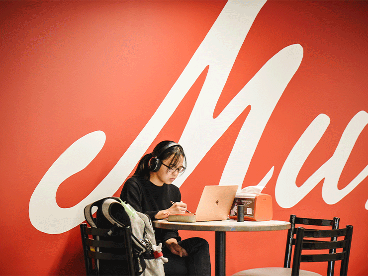 A young adult sits at a table in a cafe area, wearing headphones and working from a laptop. The wall behind them includes a large cursive 