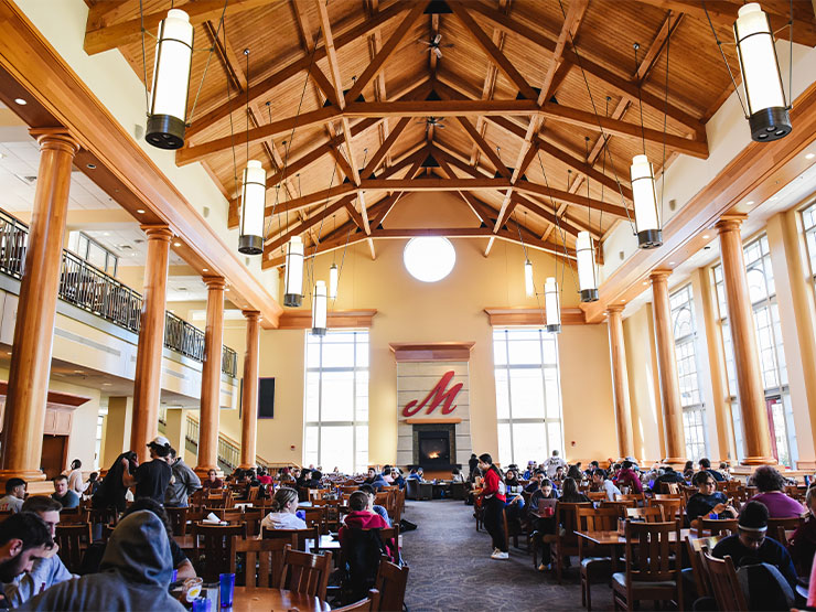 Students pack Wood Dining Hall, a large room with a high ceiling and fireplace against a far wall with scripted 