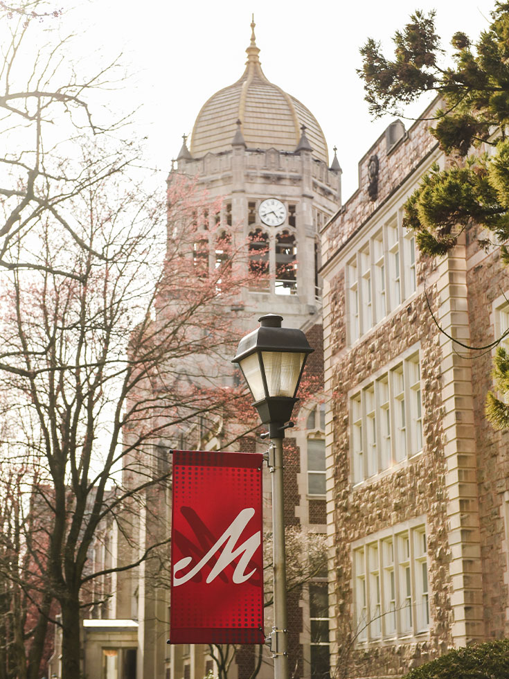 The Haas College Center clocktower stands against a grey sky with trees starting to bud and a red banner with a scripted M hanging from a light pole below.