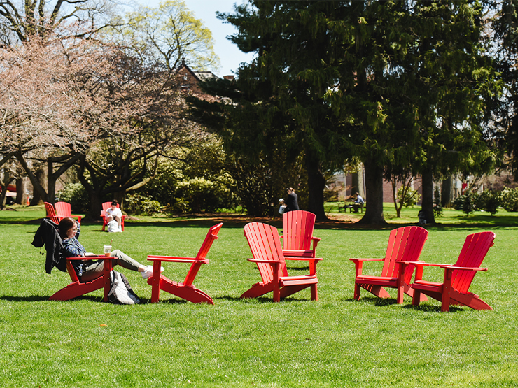 A student sits in one of a cluster of red Adirondak chairs gathered on the Muhlenberg College green.