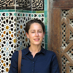 A woman in a dark blue blouse smiles at the camera while standing in front of a wall of colorful, elaborately arranged tiles.