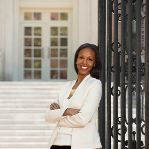 A smiling person in a white blazer leans against a black wrought iron fence.