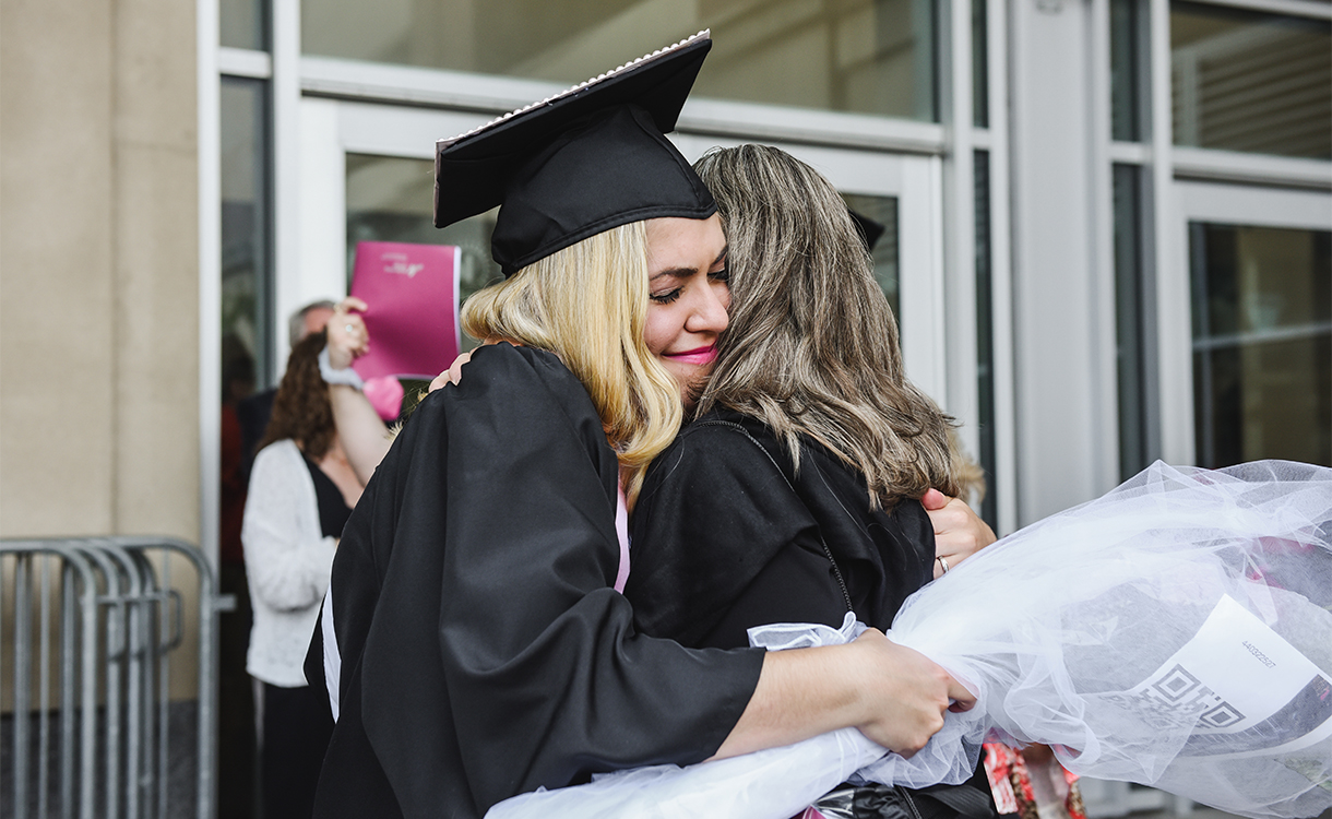 A student, dressed in a cap and gown and clutching flowers, embraces a woman.