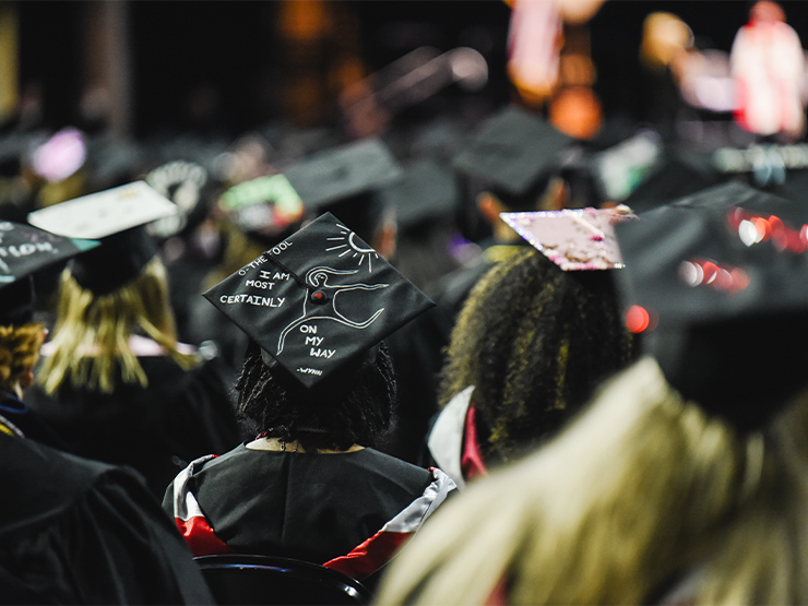 Pictured from behind, students wearing decorated graduation caps look toward a distance stage.