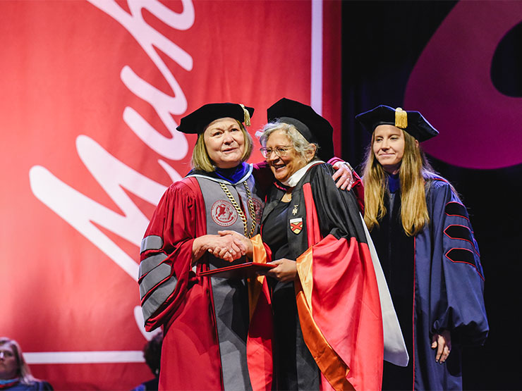 Three women in various commencement regalia stand on a stage together, two of them embracing.