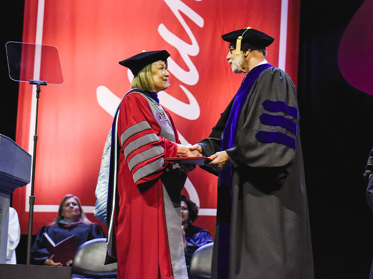A man and woman in graduation regalia shake hands, smiling, as a degree is exchnaged.