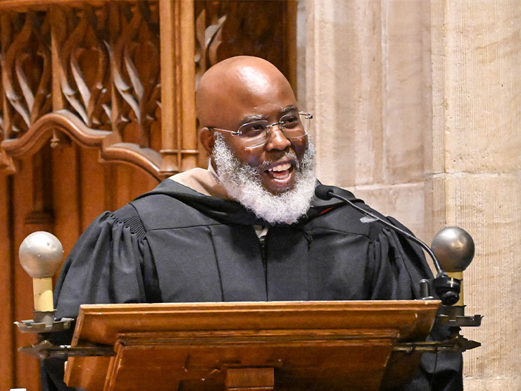 A man with greying beard speaks at a podium in a chapel while wearing a graduation robe.