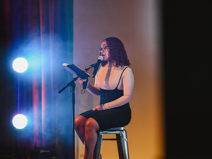 A young adult with curly, shoulder-length hair, reads from a notebook while sitting on a stool on a stage with dramatic lighting.