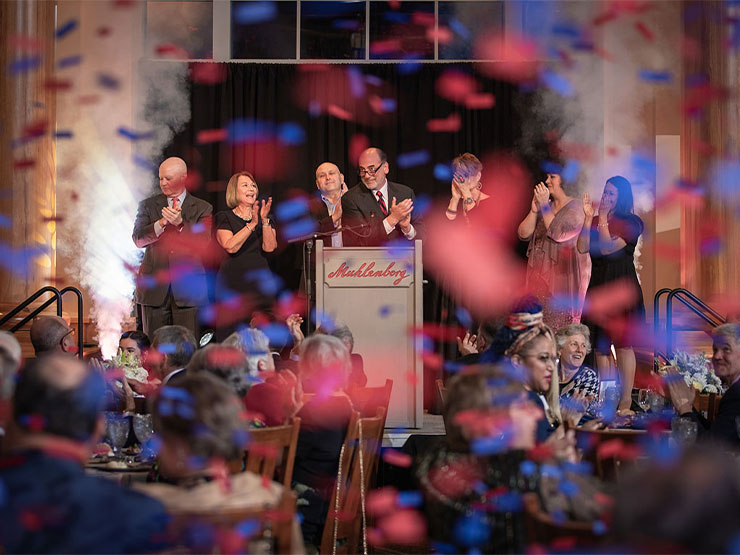 A group of people applaud on a stage in front of a crowd as blue and red confetti rains down.