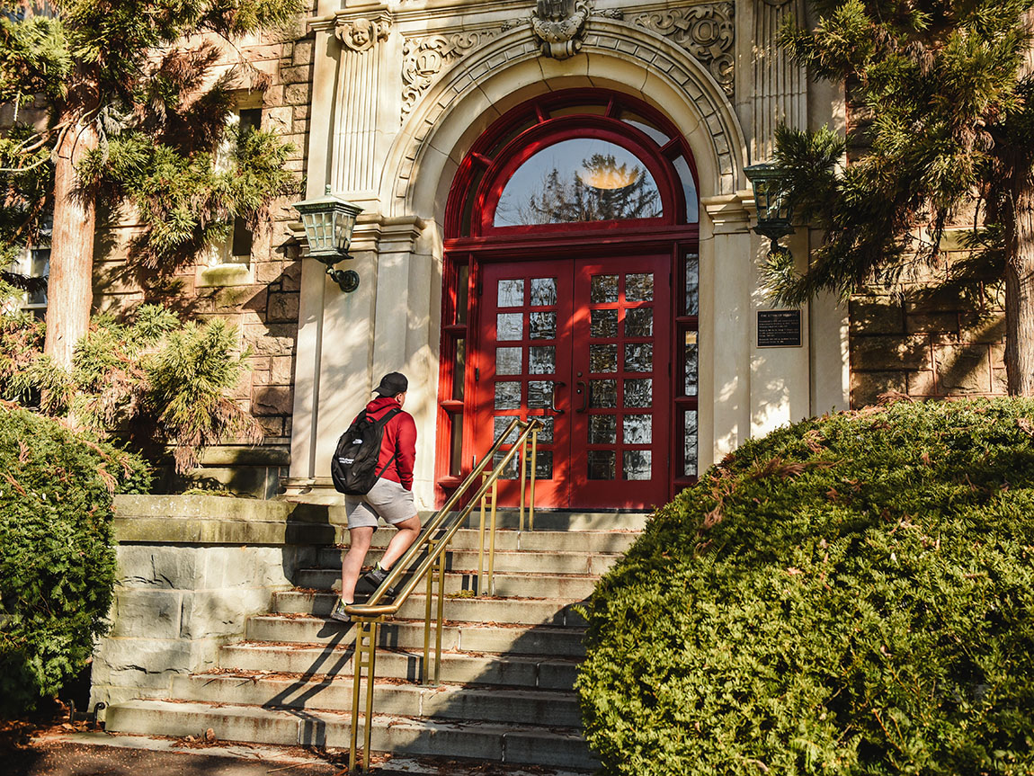 A college student goes up stairs toward a pair of red doors on the exterior of a building