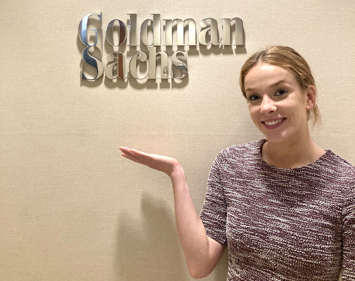 Adrienne standing in the Goldman Sachs office in Philadelphia, wearing a maroon and white dress and looking very happy with a Goldman Sachs sign behind her