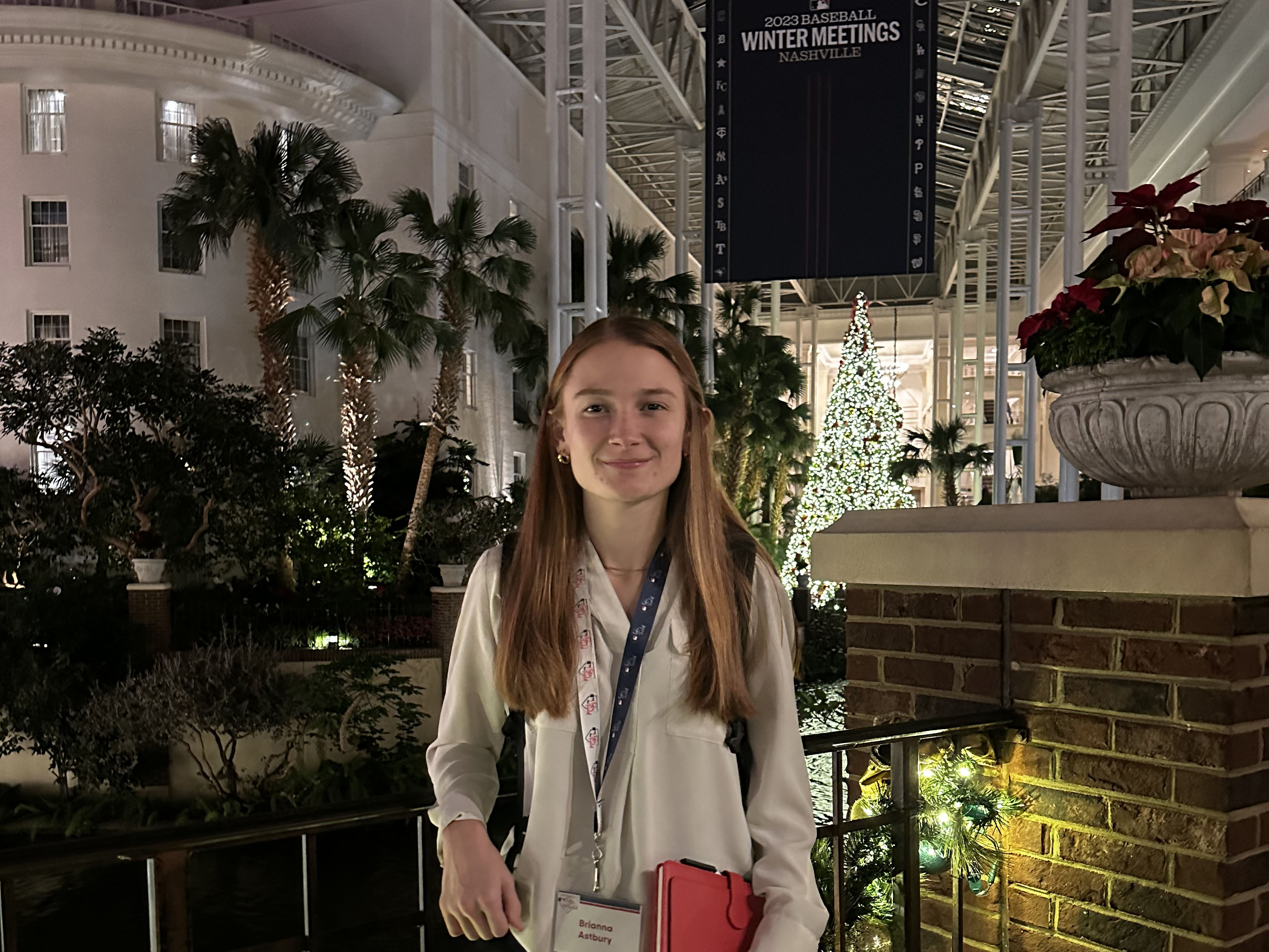 A college student stands in front of a Christmas tree and palm trees and a sign that says 2023 Baseball Winter Meetings Nashville