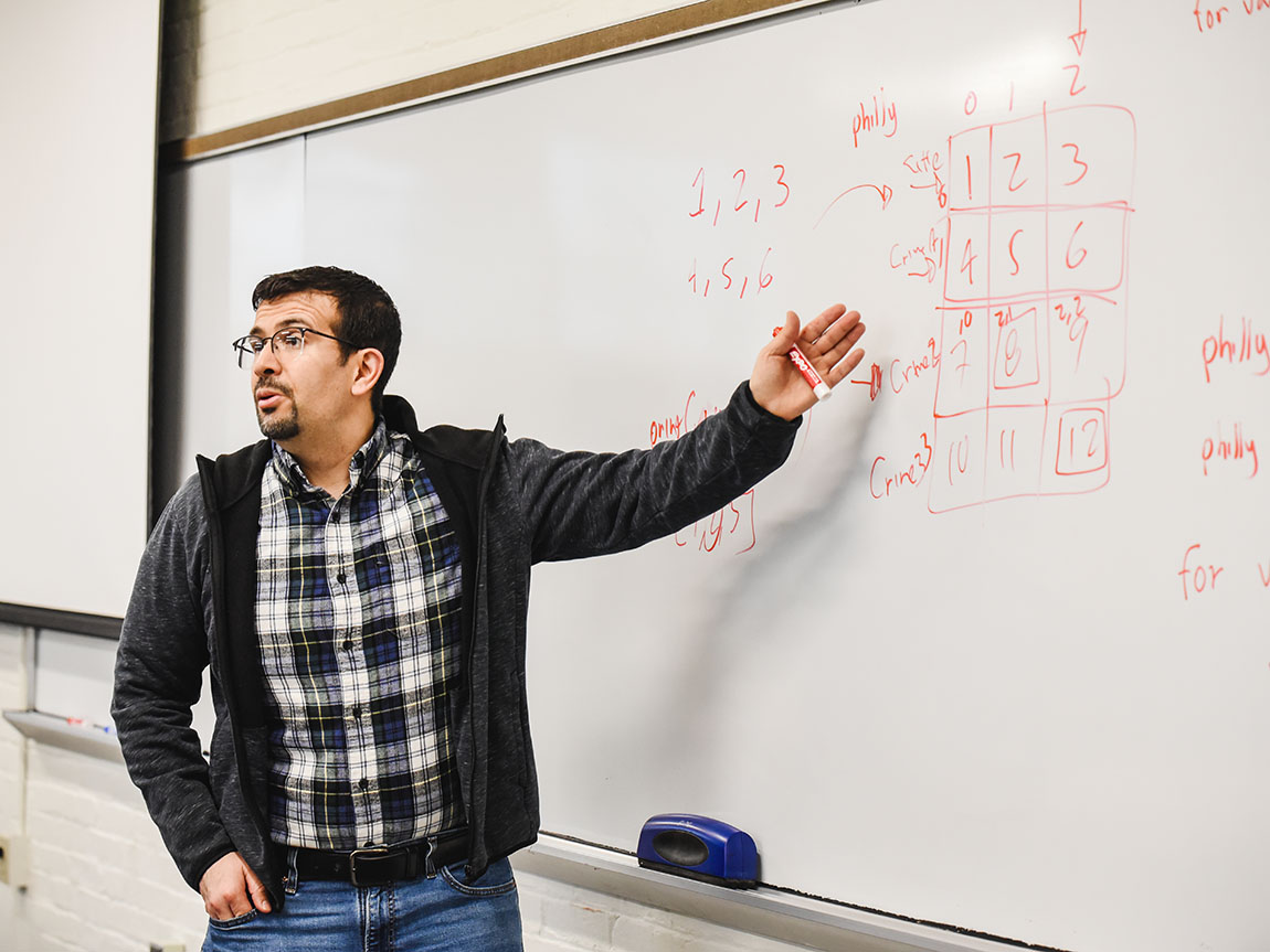 A college professor wearing glasses and a plaid shirt points at a whiteboard