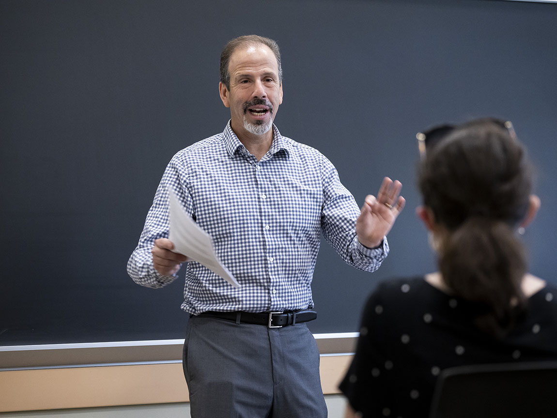 A college professor speaks to his students at the front of a classroom in front of a chalkboard