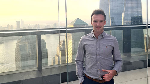 A recent college graduate in a button-down shirt stands on a balcony with a city skyline in the background.