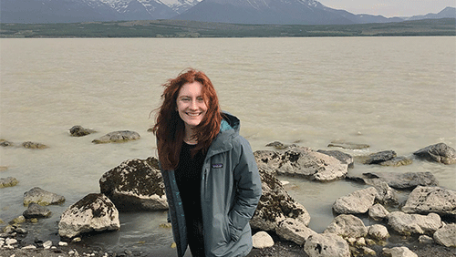 A young adult with wavy red hair smiles at the camera on the rocky shore of a lake.
