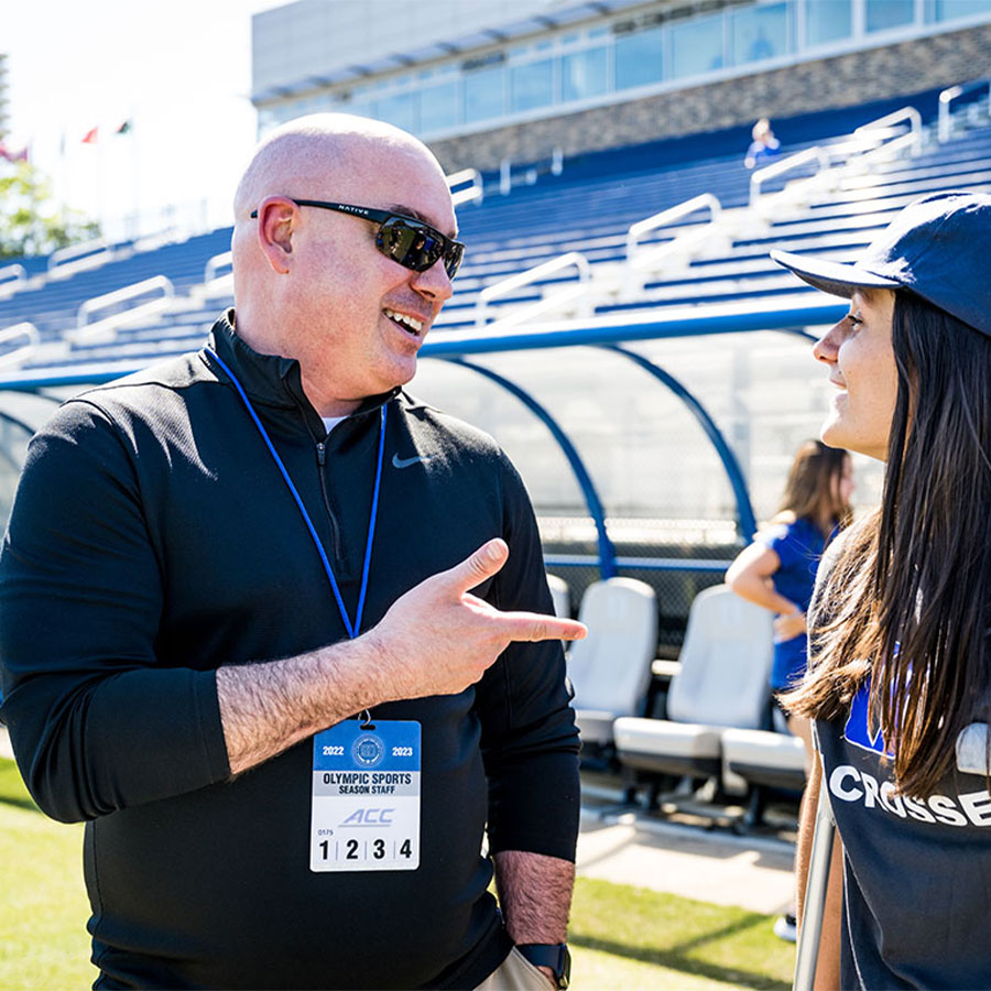 A bald man in sunglasses speaks with a college student with long hair wearing a baseball cap and using crutches with bleachers in the background