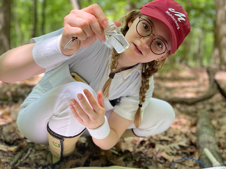 A research student wearing a red Muhlenberg College ball cap crouches on a leaf-covered forest floor, examining a vial.