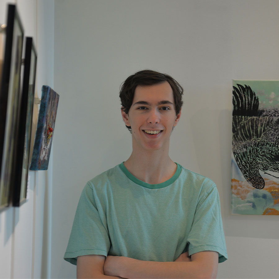 A young adult in a green tee-shirt stands with his arms crossed, smiling at the camera in the room of an art gallery.