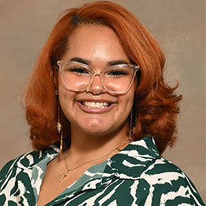 A photo of headshot of a woman with short red hair and glasses wearing a green and white patterned blouse.