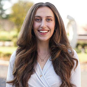 A woman with long brown hair wears a white blouse and smiles outdoors.