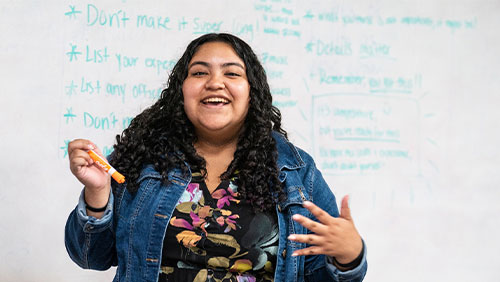 A young adult with long, curly black hair and a denim jacket laughs in front of a whiteboard full of notes.